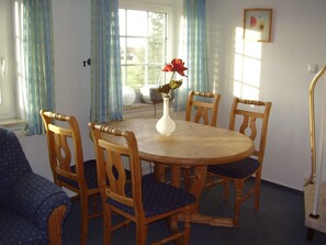 Un coin repas avec une table ronde en bois, quatre chaises en bois avec des coussins bleus, un vase blanc contenant une fleur rouge et une fenêtre ornée de rideaux bleus.