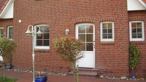 A red brick building with a white door and windows, a bell, and a small tree in the foreground. - Vacation home in Dornumersiel 80-142b - 800-142b (Dornum)