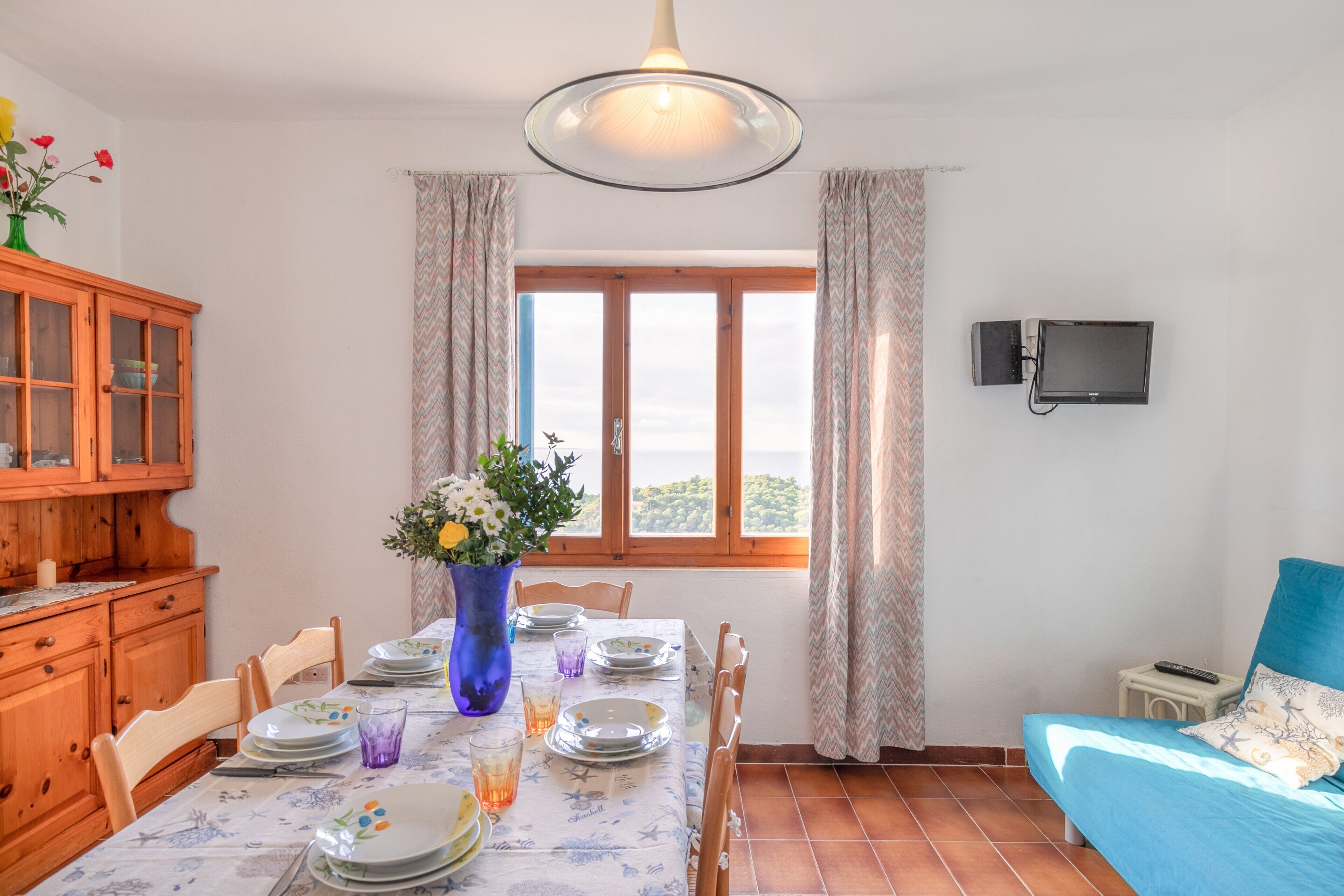 Un coin repas avec une table dressée pour quatre personnes, un buffet en bois, un téléviseur et un lit recouvert d’une couverture bleue.