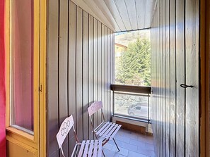 A wooden balcony with a bench and chairs, a stone wall, and a view of trees and a parked car. - Economical studio for 2 in Reberty 1850, near slopes (Les Menuires)