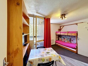A room with a bunk bed, a table with a fruit-patterned tablecloth, and a window with red curtains.