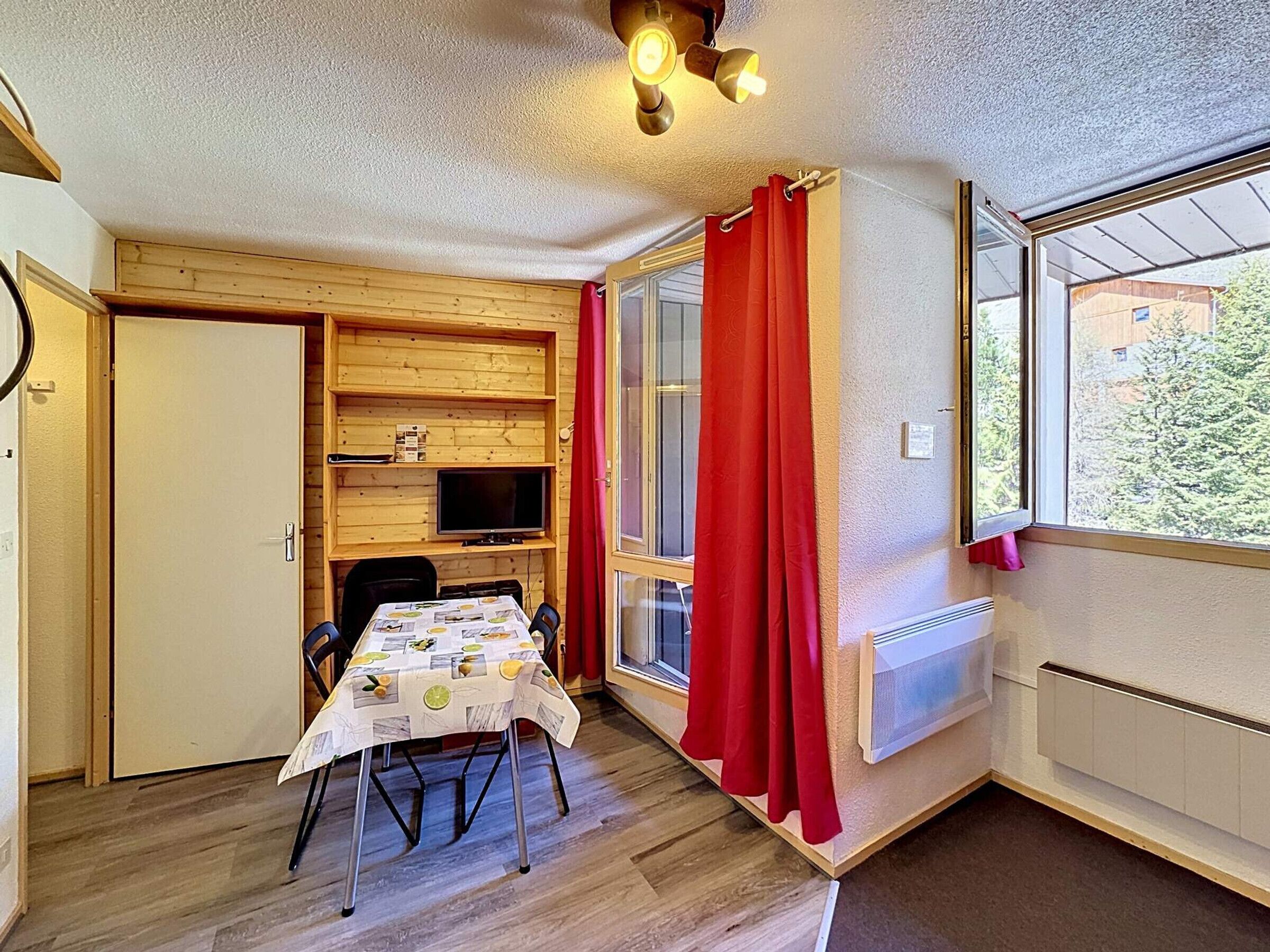 Quarto compacto com uma mesa de jantar, cadeiras, televisão e uma janela com cortinas vermelhas.