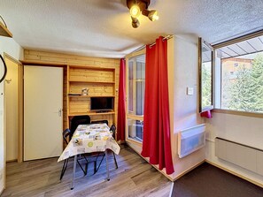 A compact room with a dining table, chairs, a television, and a window with red curtains.