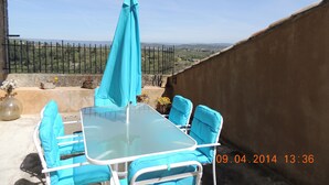 Una terraza con una mesa de vidrio y sillas azules, un paraguas turquesa, y vista a una cerca y un paisaje lejano.