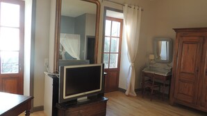 Un baño con tocador de madera, un espejo, un televisor sobre una cómoda y una ventana con cortinas.