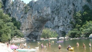 Formación rocosa natural con una abertura parecida a una cueva, rodeada de vegetación y una masa de agua donde la gente participa en actividades acuáticas.