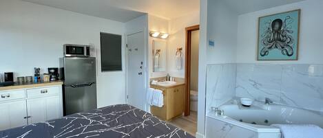 Una habitación de hotel compacta con cama, kitchenette, baño y un cuadro en la pared.