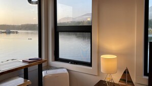 A room with a large window offering a view of a lake and mountains, a desk with a lamp, and a small heater.
