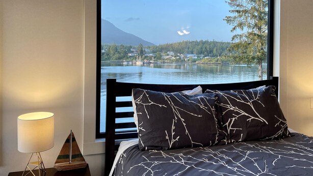 A bedroom with a large window overlooking a lake, a bed with patterned bedding, and a bedside lamp.