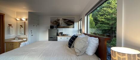 A bedroom with a large bed, a bedside table, a lamp, a window with a view of trees, a kitchen area with a refrigerator and microwave, and a bathroom with a sink and mirror.