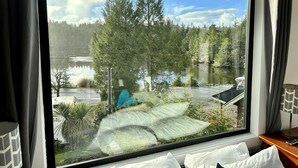 A large window with a view of a lake, trees, and a building with a blue tarp. - Sea Esta (Ucluelet)