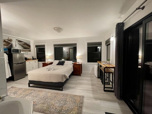 Ein kleines Hotelzimmer mit Bett, Nachttischen, einer kompakten Küchenzeile und einer Glasschiebetür, die auf einen Balkon führt.