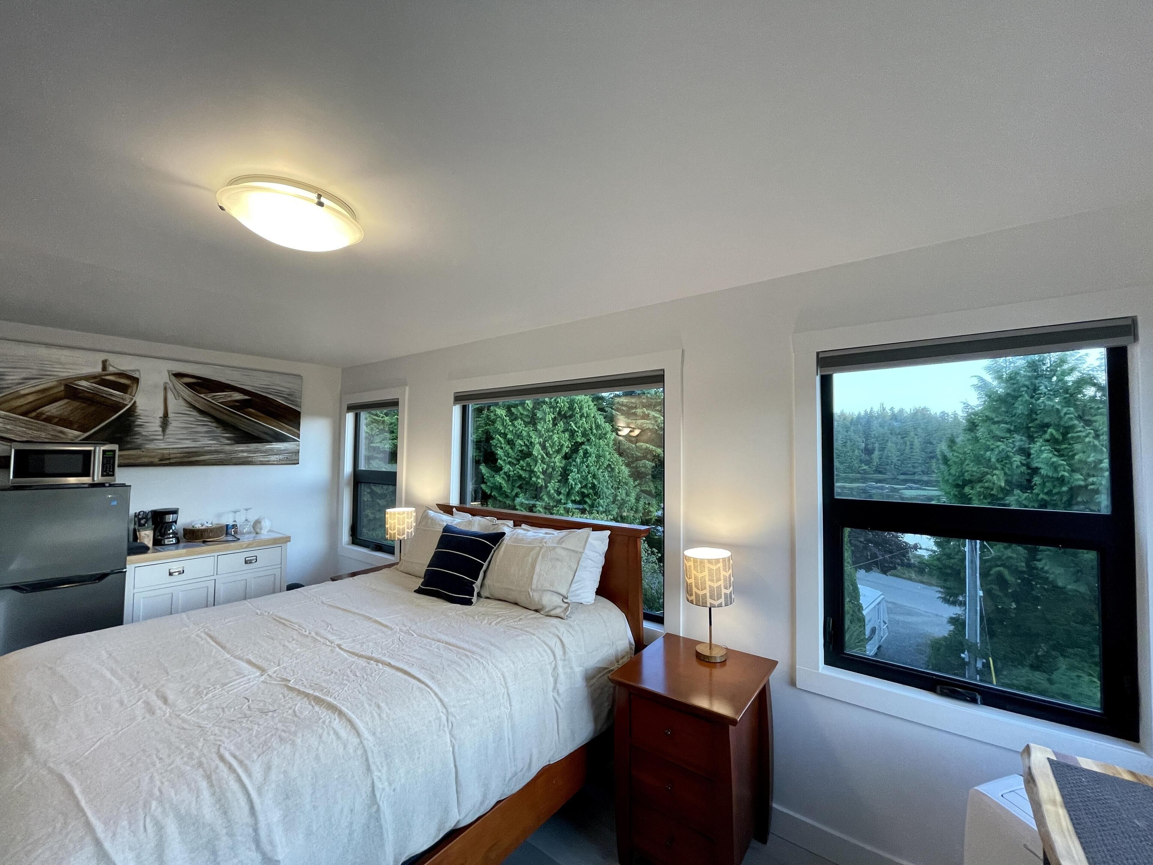 A bedroom with a bed, bedside tables, a lamp, a microwave, and a refrigerator. There are large windows with a view of trees and a building.