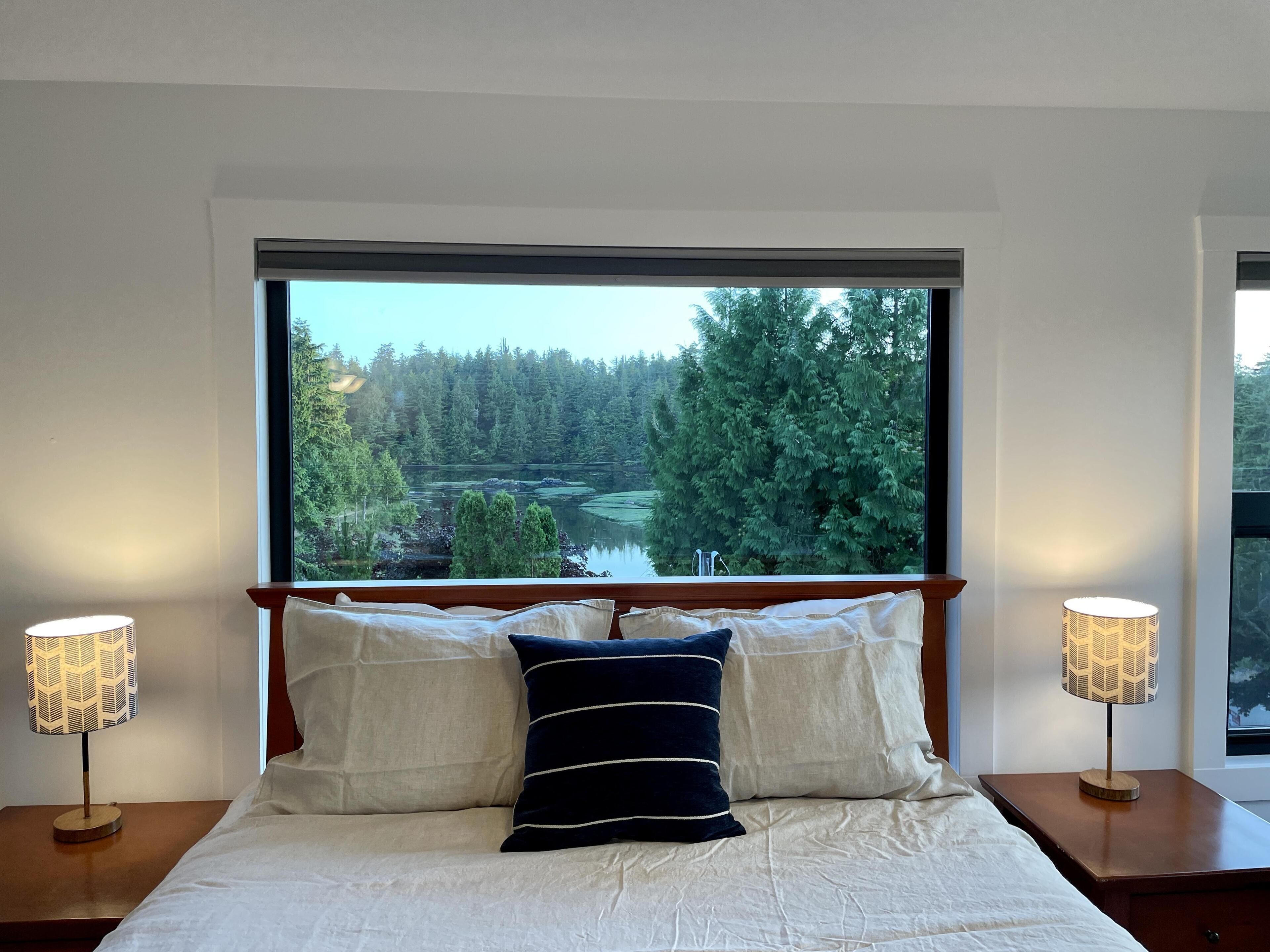 Une chambre à coucher dotée d’une grande fenêtre donnant sur une forêt et un lac. On y trouve deux tables de chevet avec des lampes.