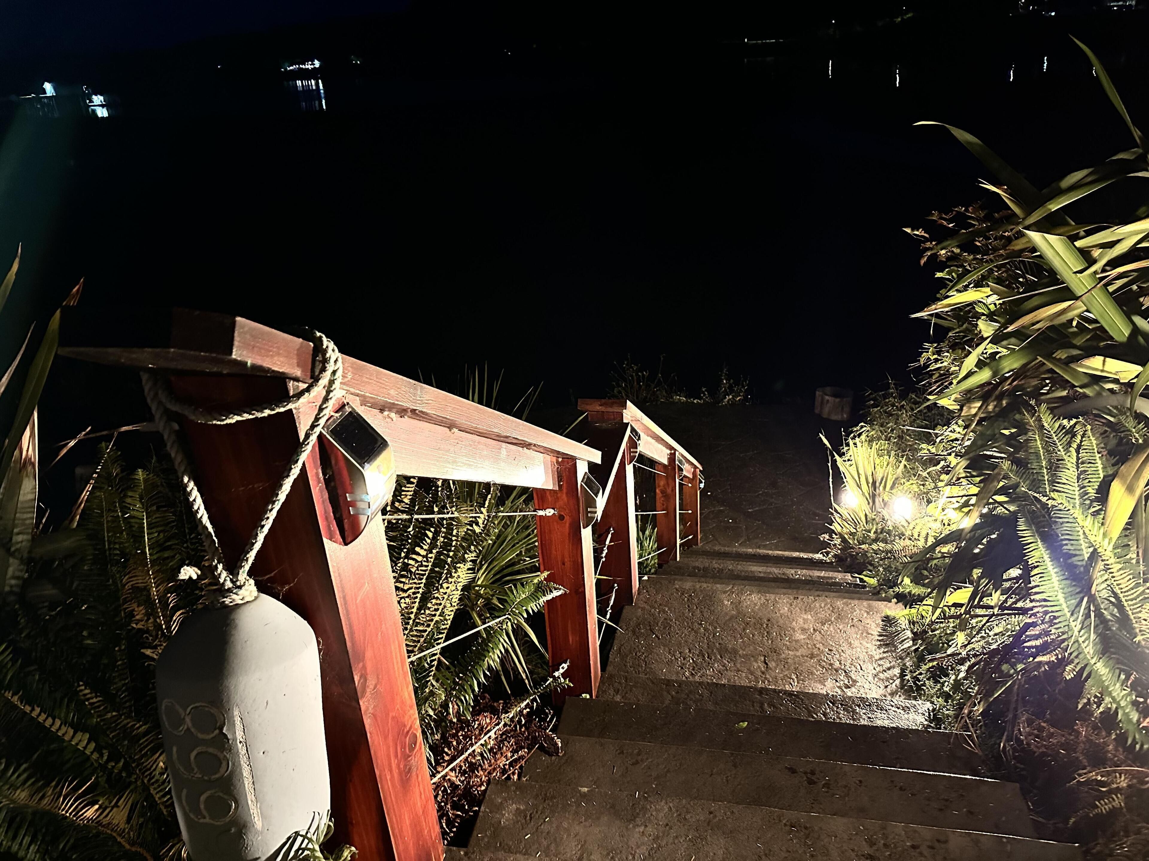 A wooden bridge with rope railings, surrounded by greenery and illuminated by artificial lighting.