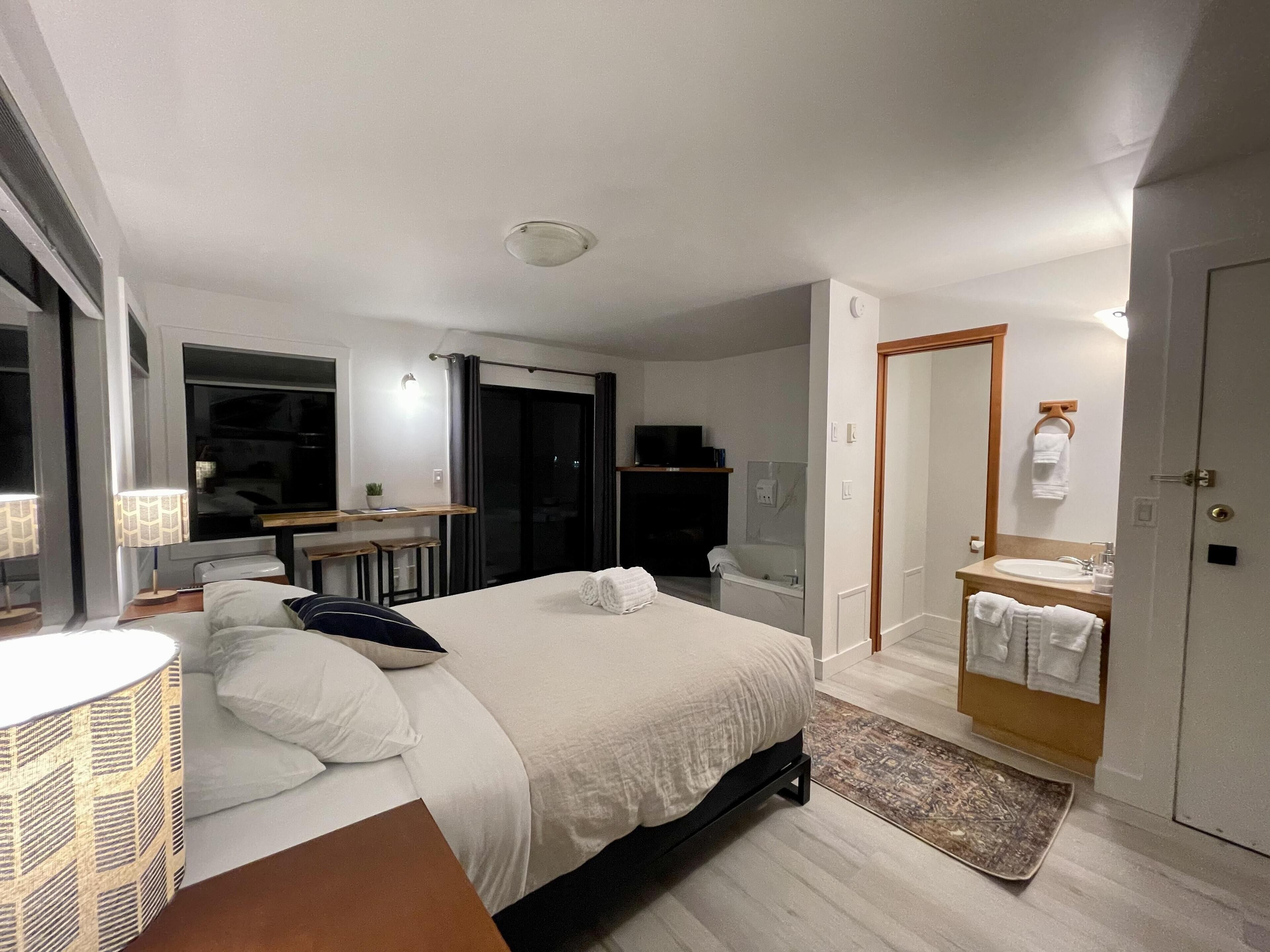 Ett modernt hotellrum med en säng, ett nattduksbord, en lampa och utsikt över staden på natten.