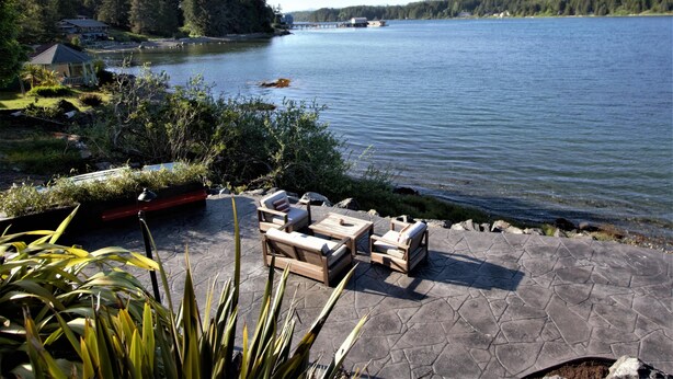 A lakeside patio with outdoor furniture, a dock, and a house in the background. - Sea Esta (Ucluelet)