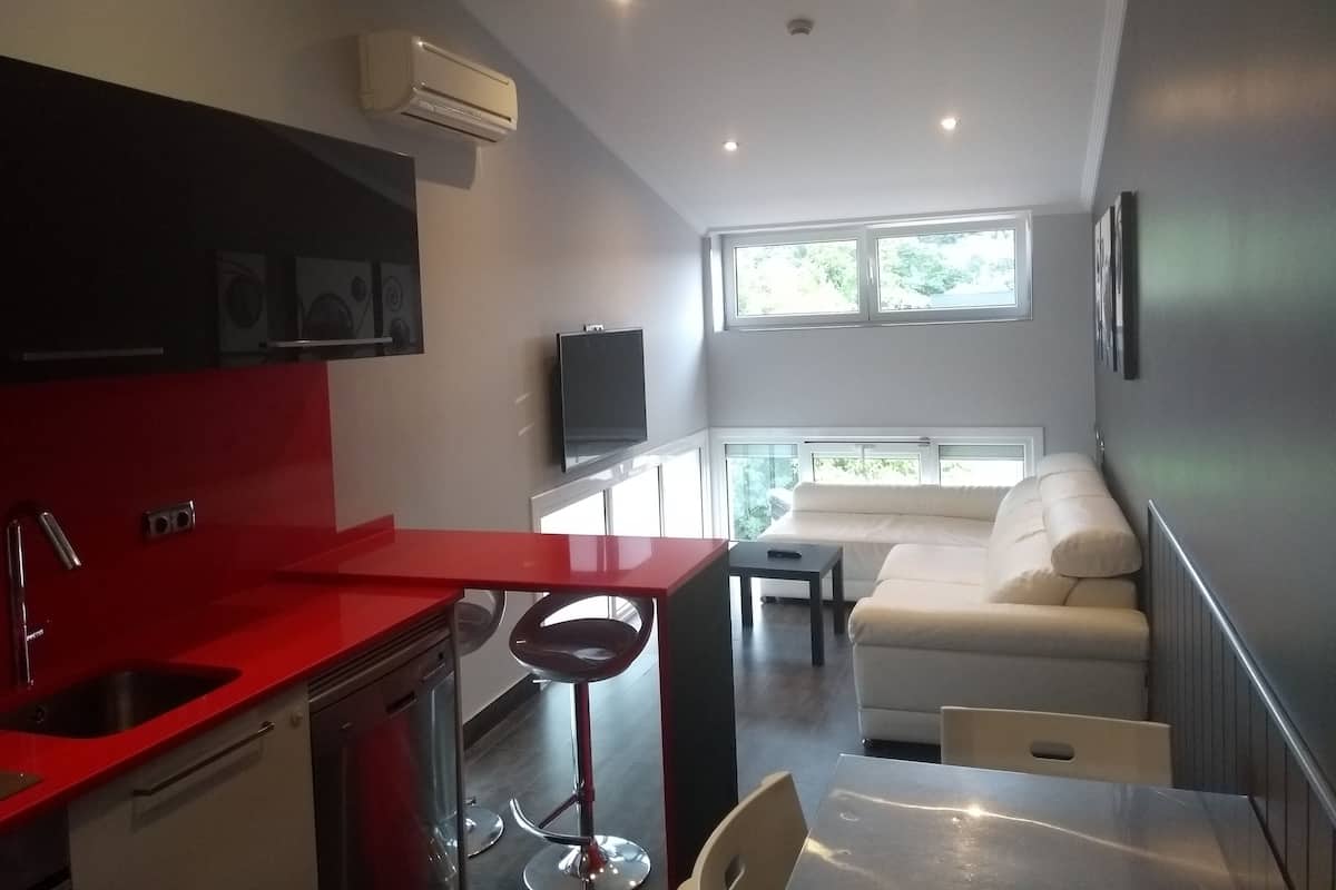 A compact kitchen with a red countertop, a built-in stove, and a sink. There's a bar stool, a small table with a TV, and a white sofa in a sunlit living area with large windows.