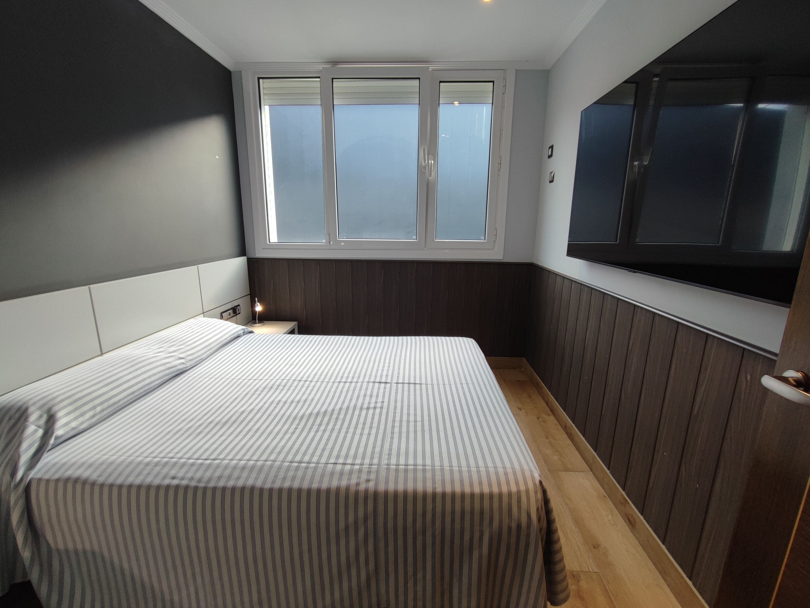 A bedroom with a bed, a nightstand, a window, and a TV mounted on the wall.