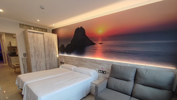 Habitación de hotel con dos camas, un sofá, un mural con atardecer sobre el agua y una kitchenette al fondo.