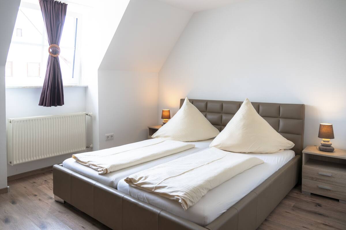 A bedroom with a large bed, two bedside tables with lamps, a window with curtains, and a radiator.