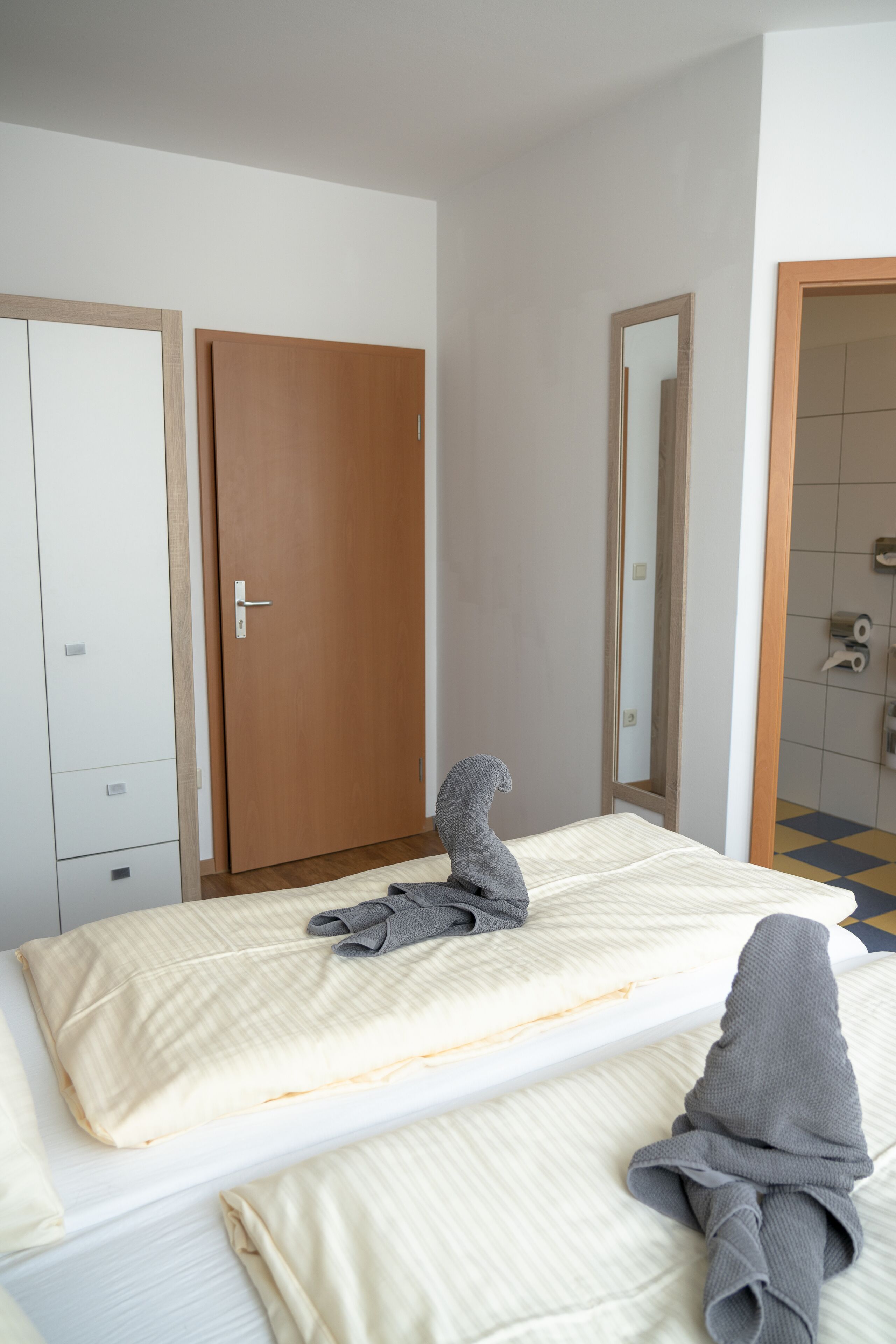 A hotel room with a bed, a wooden wardrobe, a mirror, and a bathroom with a tiled floor.