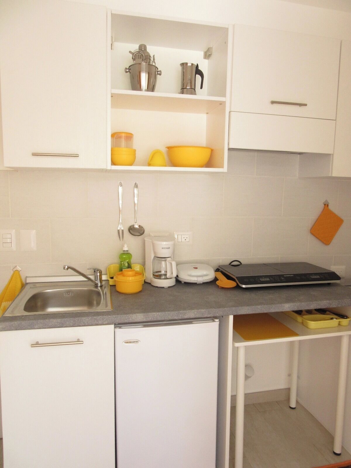 Een compacte keuken met witte kastjes, een roestvrijstalen spoelbak, een koffiezetapparaat en een broodrooster.