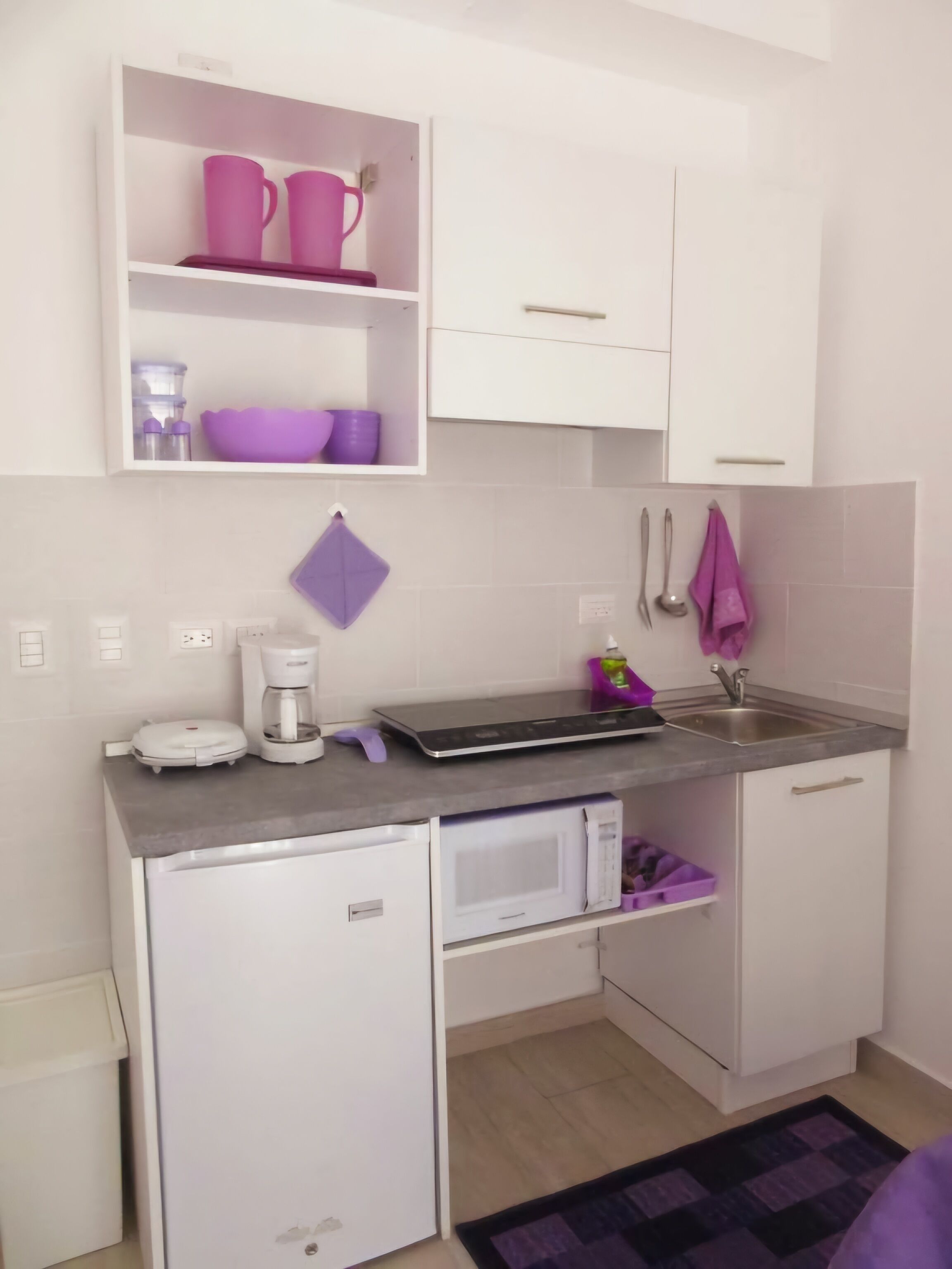 Una cocina compacta con armarios blancos, microondas, cafetera y lavamanos.