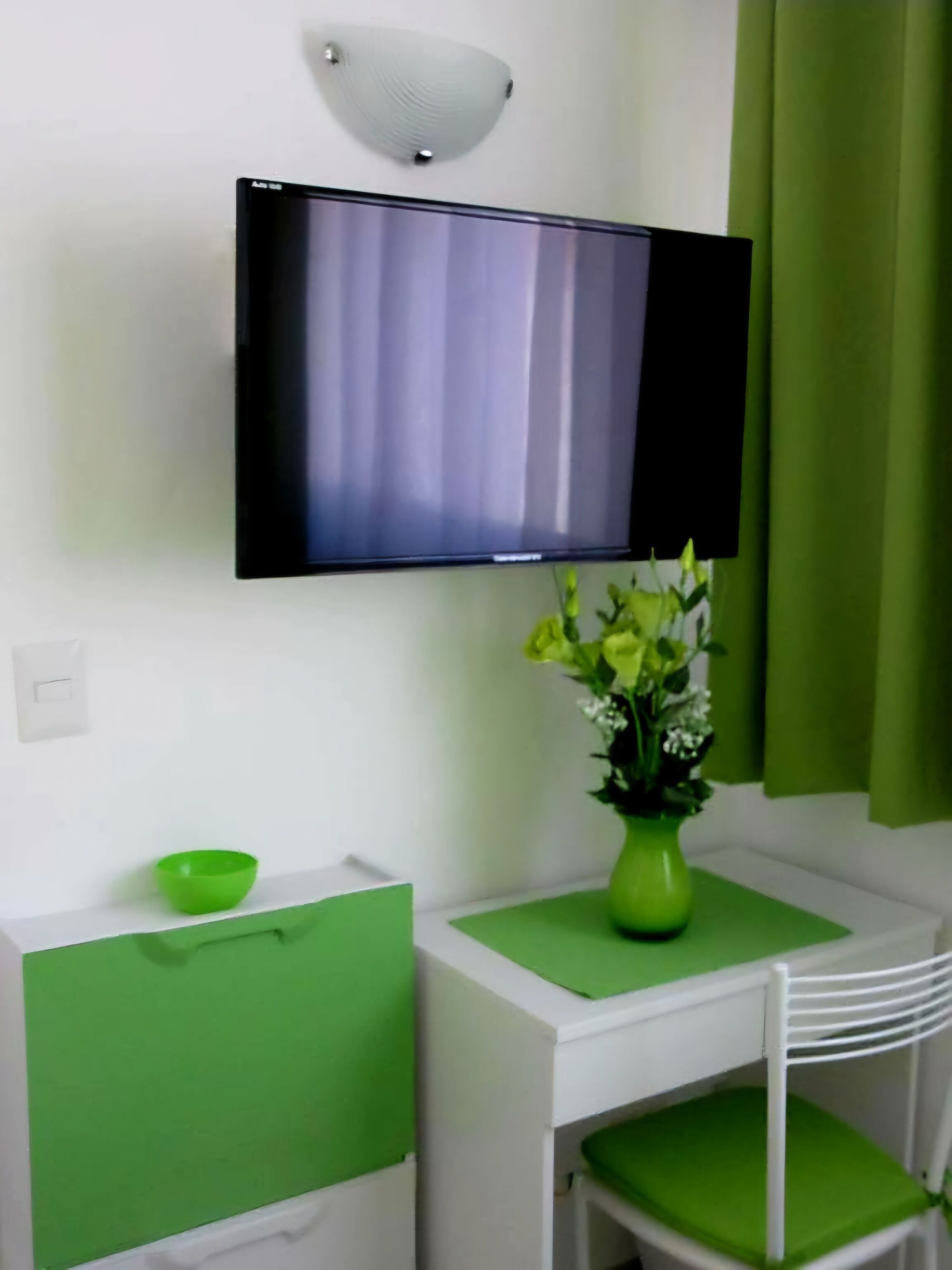A room with a flat-screen TV mounted on the wall, a green vase with flowers on a small table, a green bowl on a drawer, and a white chair with a green cushion.