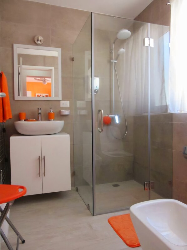 Une salle de bain moderne avec une douche à parois de verre, un lavabo blanc doté de distributeurs de savon orange et un meuble blanc avec des poignées argentées.