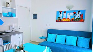 A room with a blue sofa, a dining table with chairs, a coffee machine, and a painting on the wall.