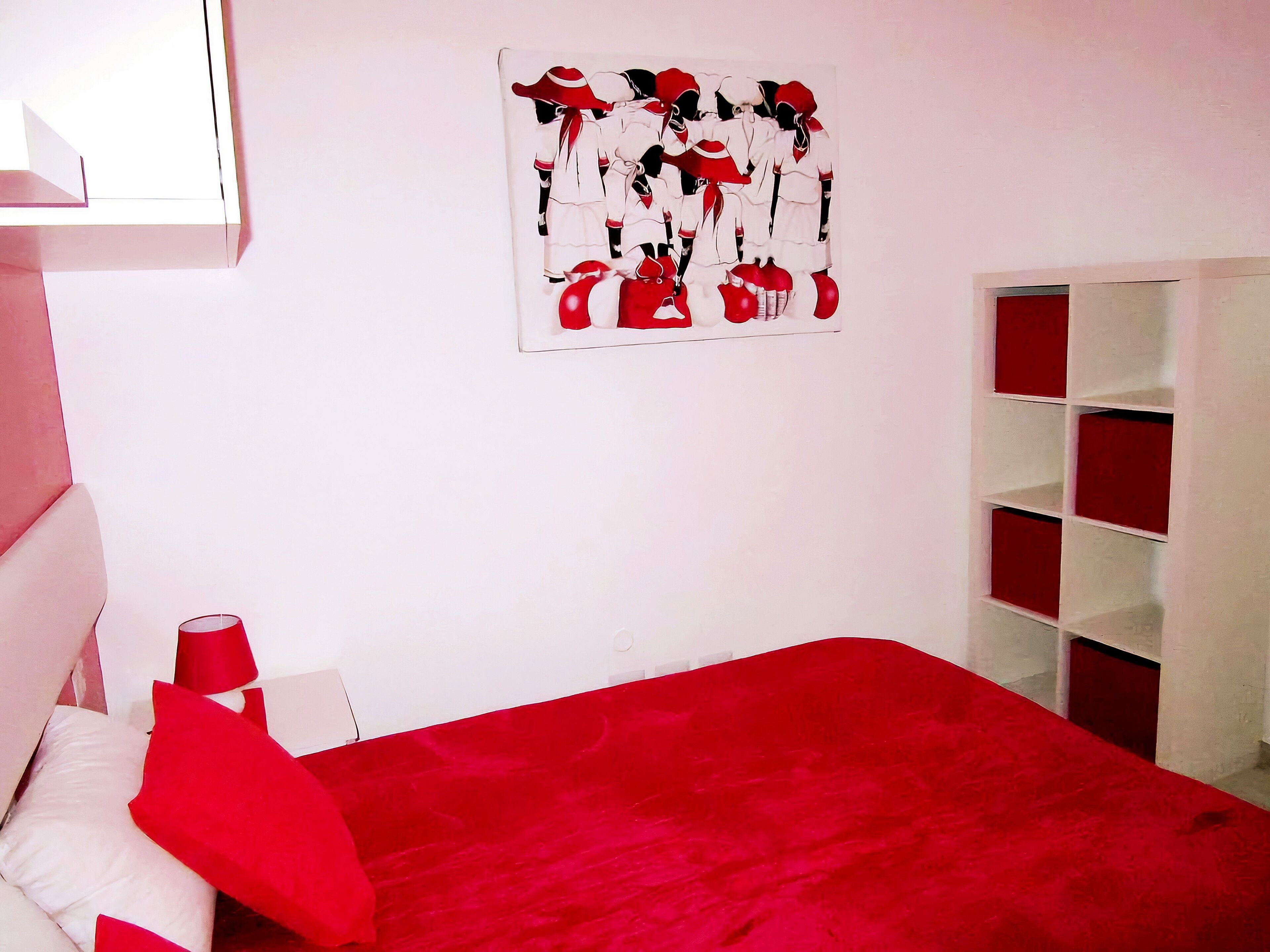 A room with a red bedspread, a white headboard, a red and white artwork on the wall, and a white shelving unit.