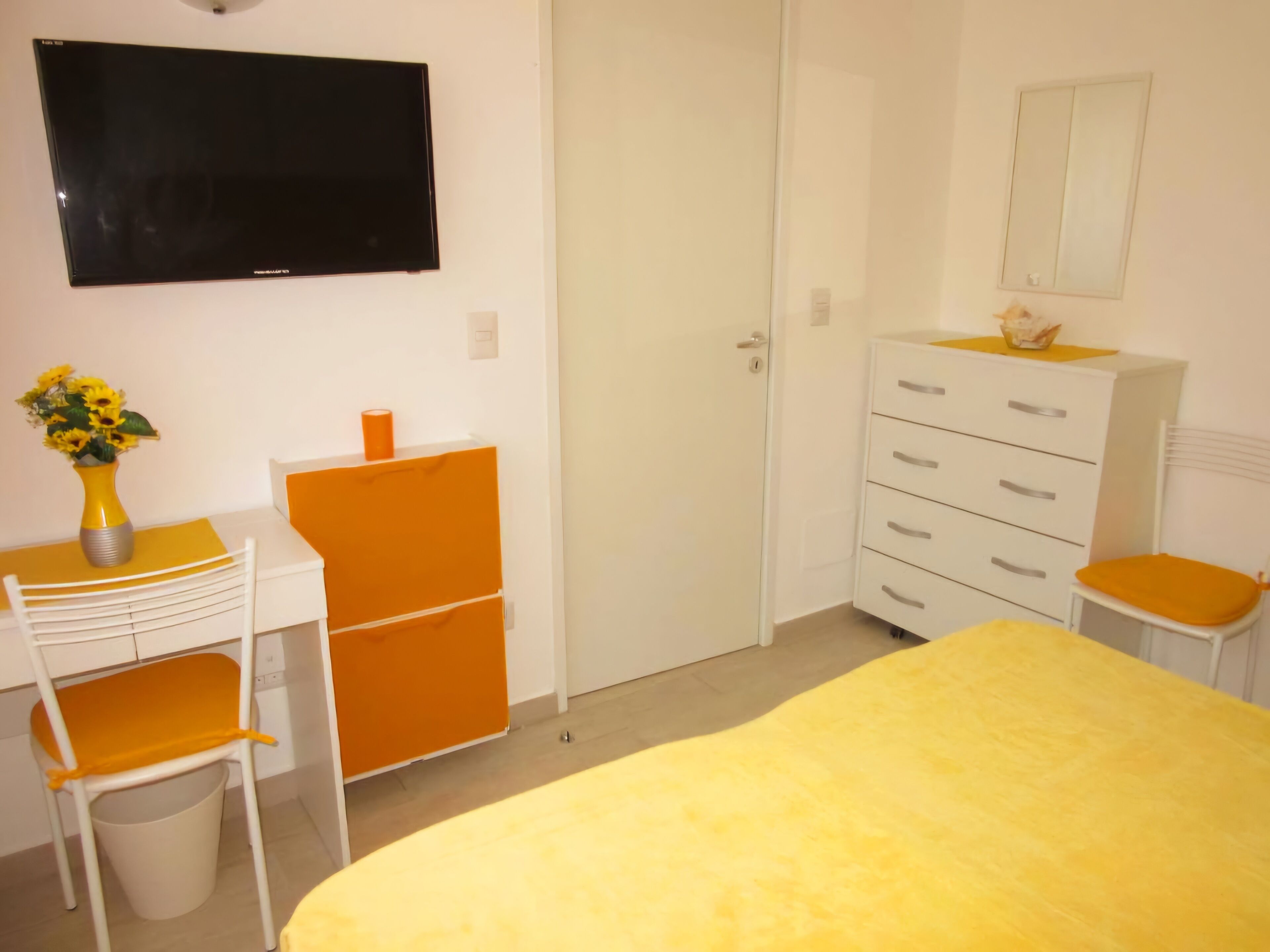 Quarto com cama, escrivaninha com cadeira, penteadeira e televisão na parede.
