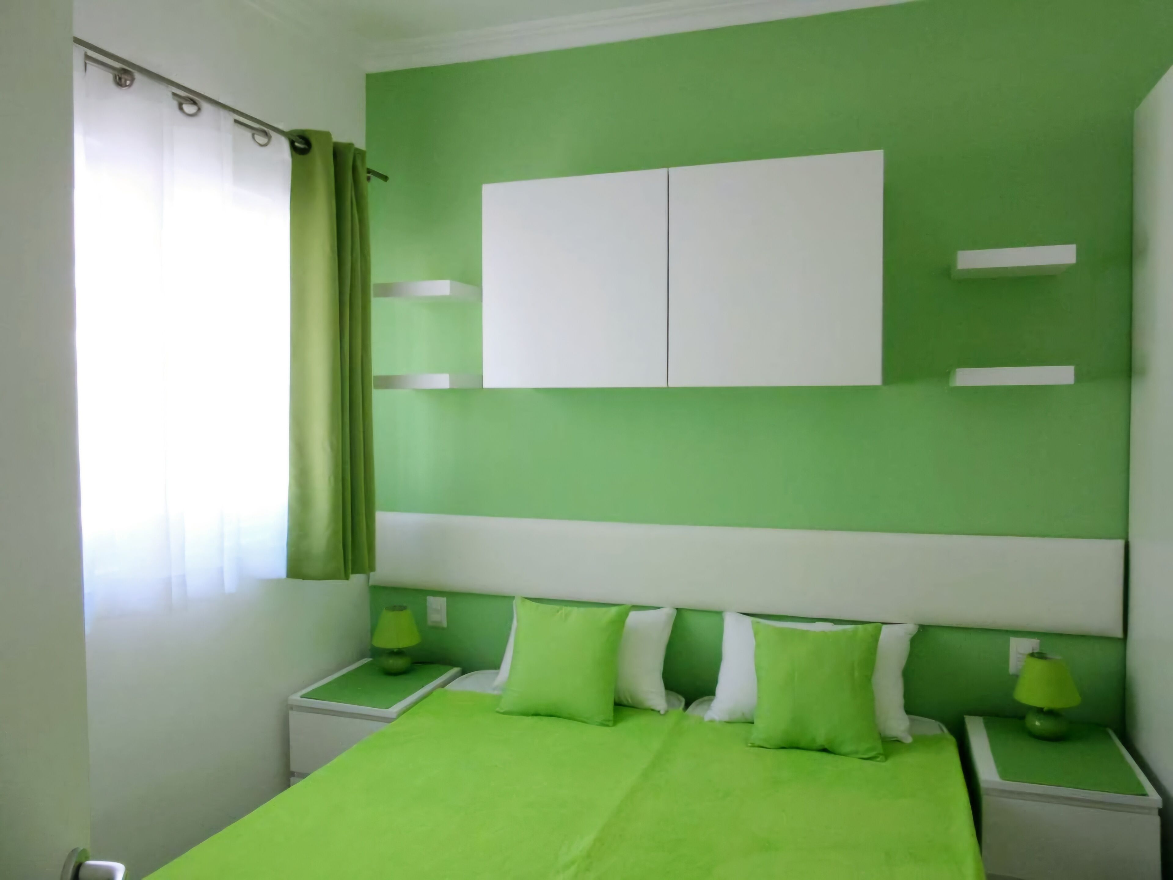 Een kamer ingericht in groen en wit, met een bed met groene beddengoed, twee nachtkastjes met lampen en een raam met een groene gordijn.