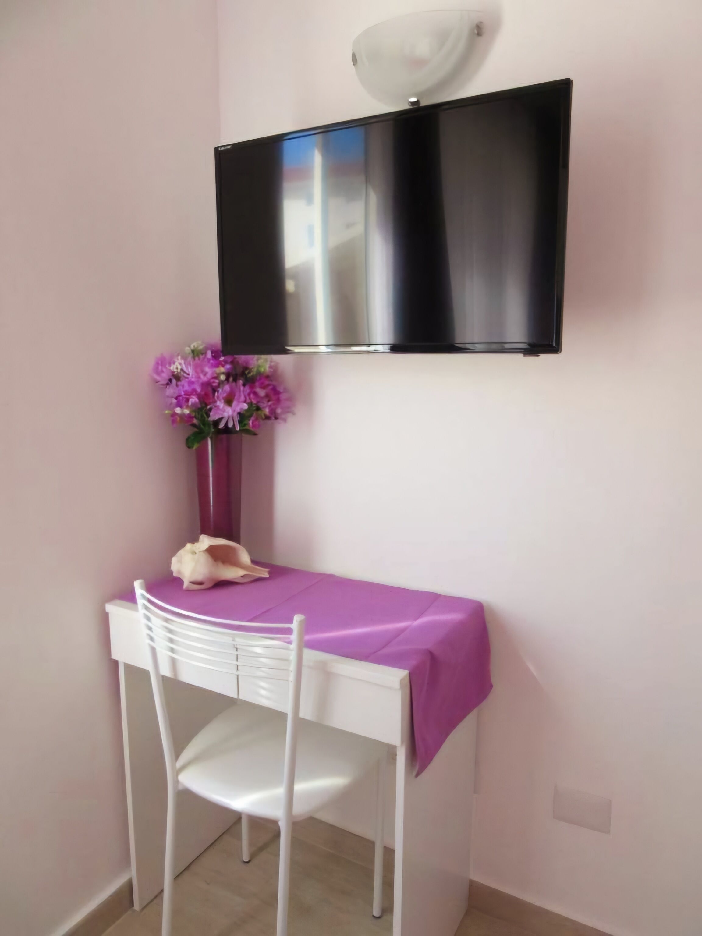 Un pequeño comedor con una mesa y silla blancos, un mantel púrpura, un jarrón con flores y un televisor de pantalla plana montado.
