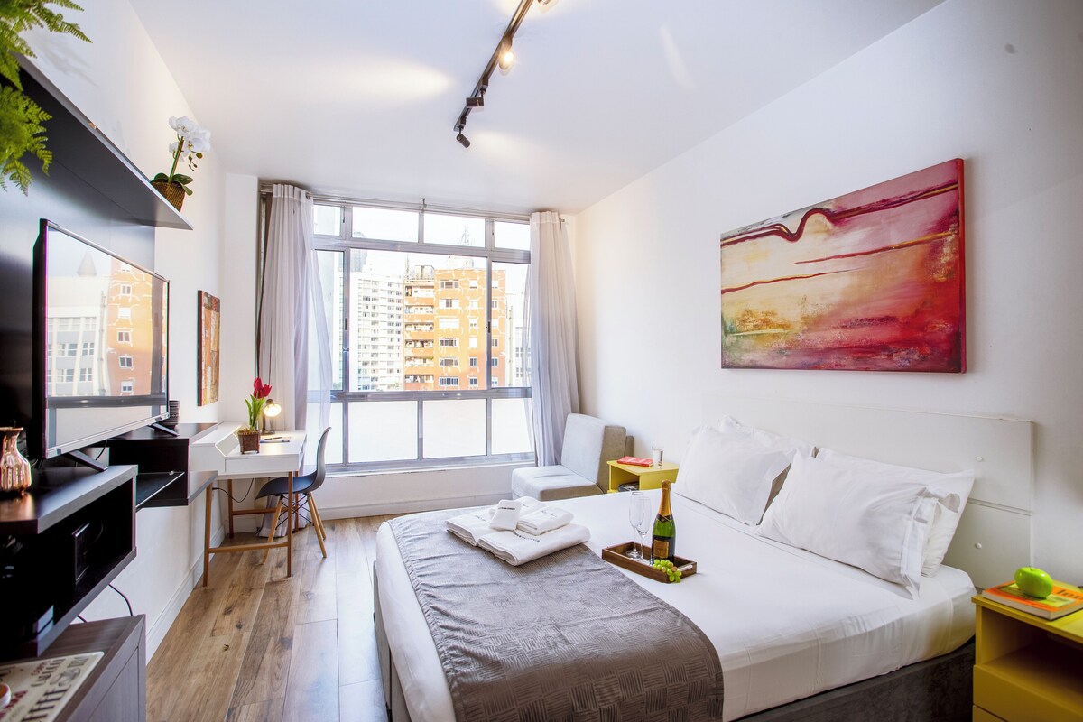Ein modernes Hotelzimmer mit einem großen Bett, einem Schreibtisch, einem Sessel, einem Fernseher und einem Fenster mit Blick auf die Stadt.
