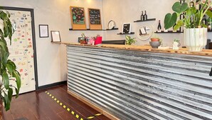 Zona de recepción con mostrador de metal corrugado, estanterías de madera con botellas y varias plantas.