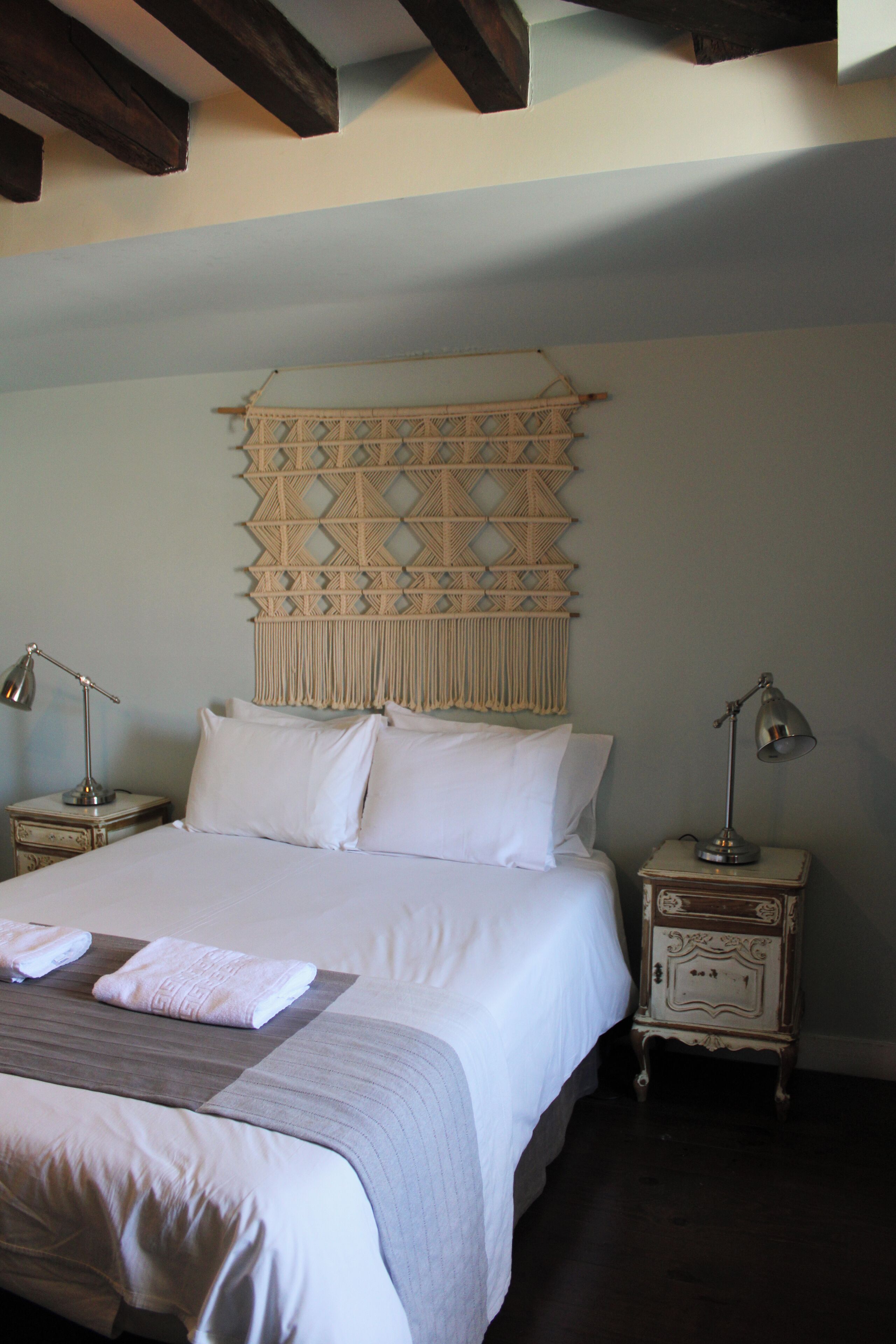 A neatly made bed with white linens and pillows, a decorative wall hanging above it, and two bedside tables with lamps.