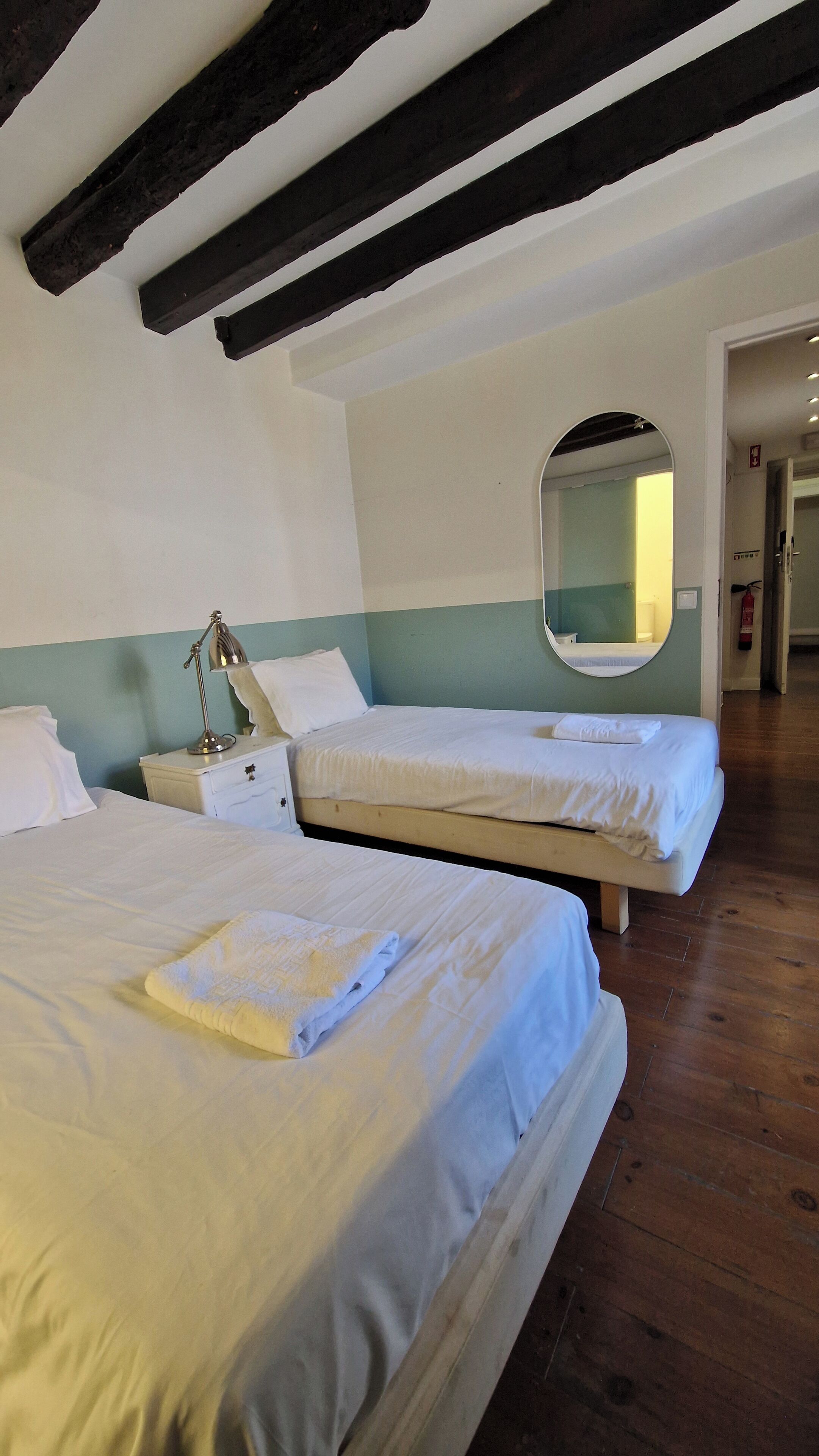 Ein Zimmer mit zwei Betten, einem Nachttisch mit Lampe, einem runden Spiegel und Holzbalken an der Decke.