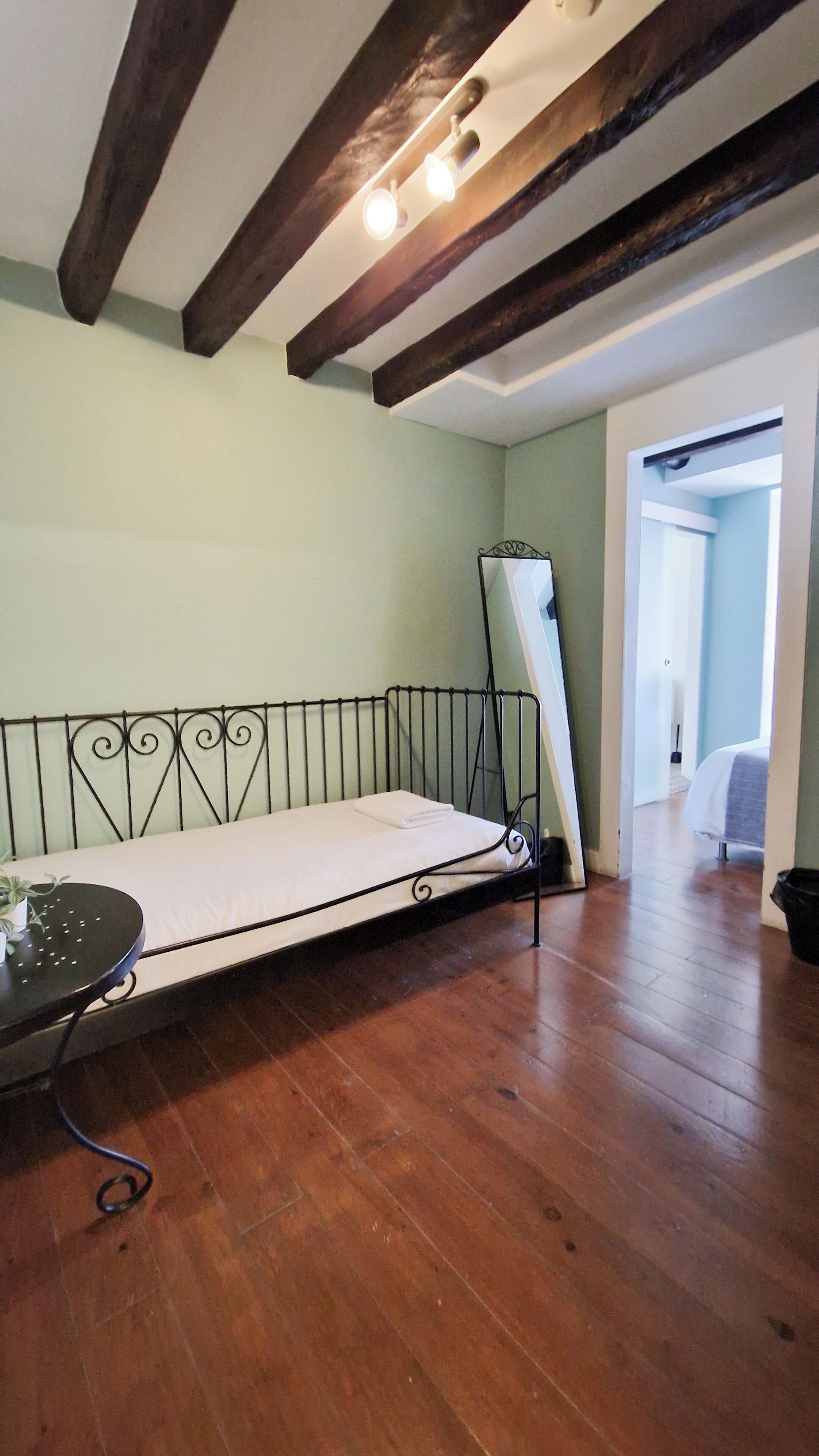 A room with a metal bed frame, a small table with a plant, wooden flooring, and exposed wooden beams on the ceiling.