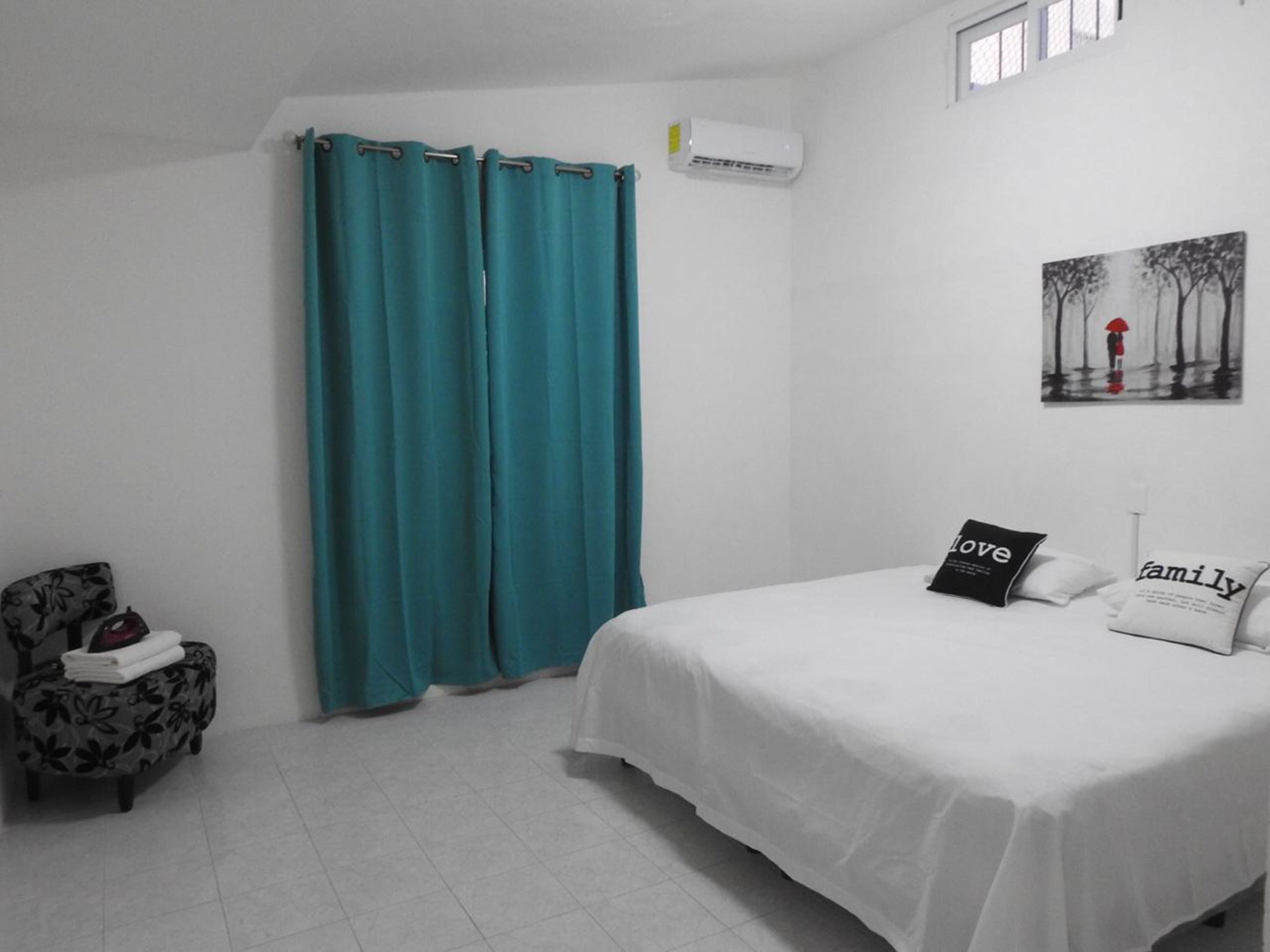 A bedroom with a bed, teal curtains, a chair with a floral pattern, and a painting on the wall.