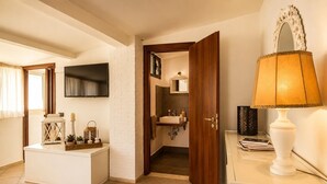 A room with a flat-screen TV, a wooden door leading to a bathroom, a bedside table with a lamp, and a white bench with decorative items. - Domus Claudia - Holiday Homes (Siracusa)