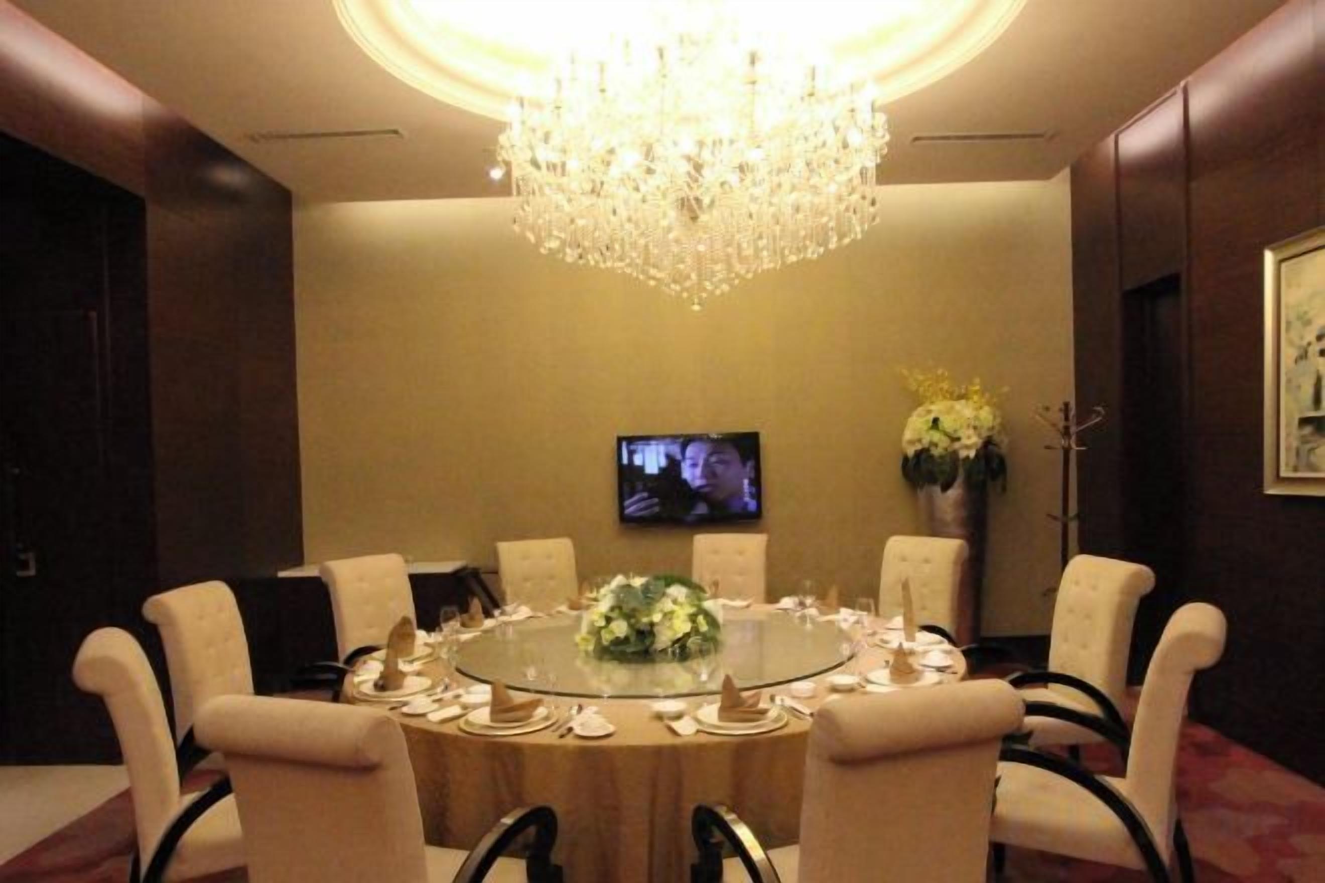 A conference room with a round table, chairs, a chandelier, a television, and a vase with flowers.