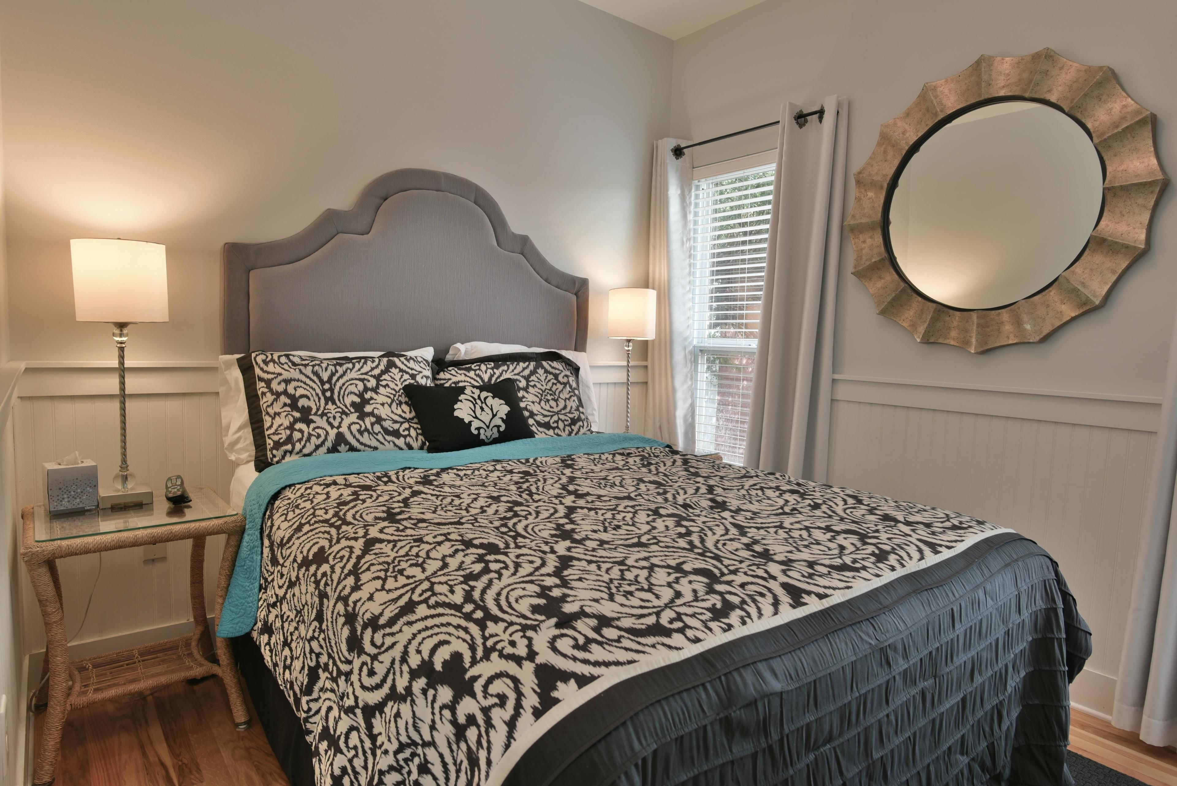 A bedroom with a bed, bedside lamps, a bedside table, a mirror, and a window with blinds.