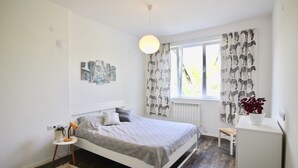 Ett sovrum med en säng, ett litet sidobord, en stol, ett element och ett fönster med mönstrade gardiner.