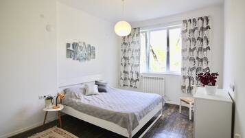 A bedroom with a bed, a small side table, a chair, a radiator, and a window with patterned curtains.