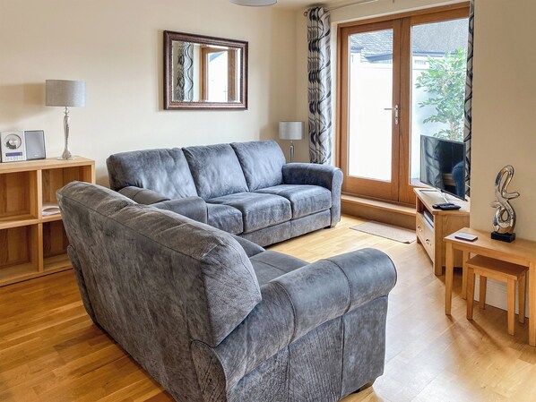 A modern living room with a grey sectional sofa, wooden flooring, a TV on a wooden stand, a lamp, and a mirror on the wall. - 2 bedroom accommodation in Keswick (Keswick)