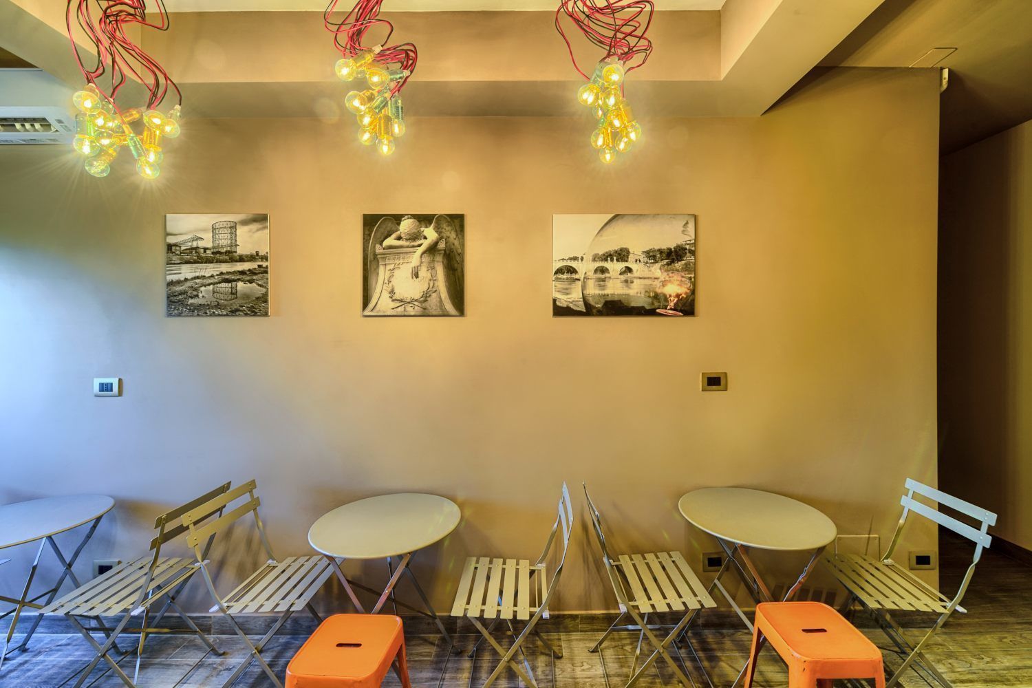 Una habitación con tres mesas redondas y sillas plegables, un candelabro en el techo y tres fotografías blanco y negro en la pared.