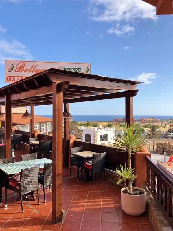 An outdoor patio area with tables and chairs, a sign for 'Bollywood Spices Curry Shop', and a view of the sea and buildings in the background. - Superb South Facing Bungalow. Great location, Air con, Wi-Fi, 2026 Samsung TV (Castillo Caleta de Fuste)