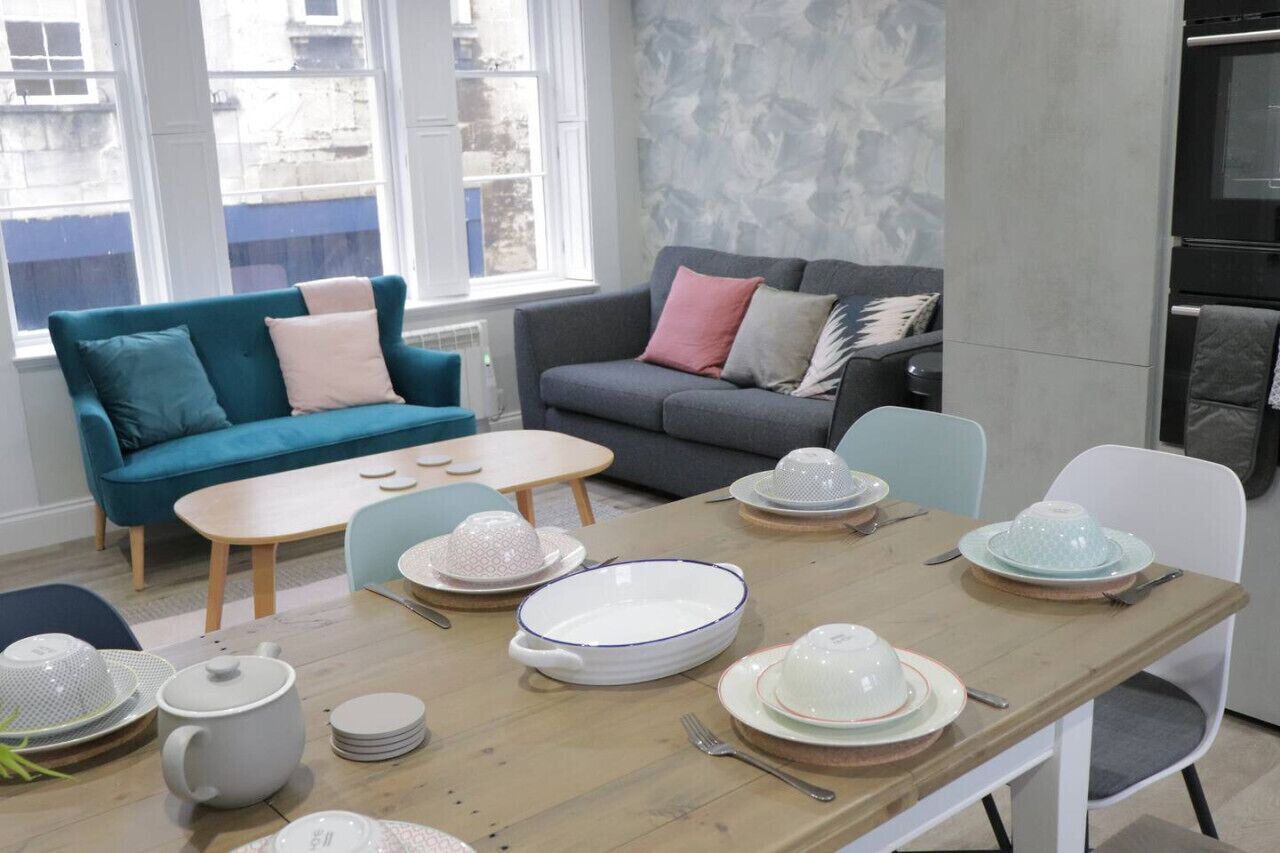 A dining area with a wooden table set for four, surrounded by a teal sofa and a grey sofa with cushions.